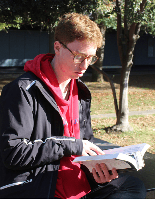 Estudiante con lentes sentando leyendo un libro en la mano